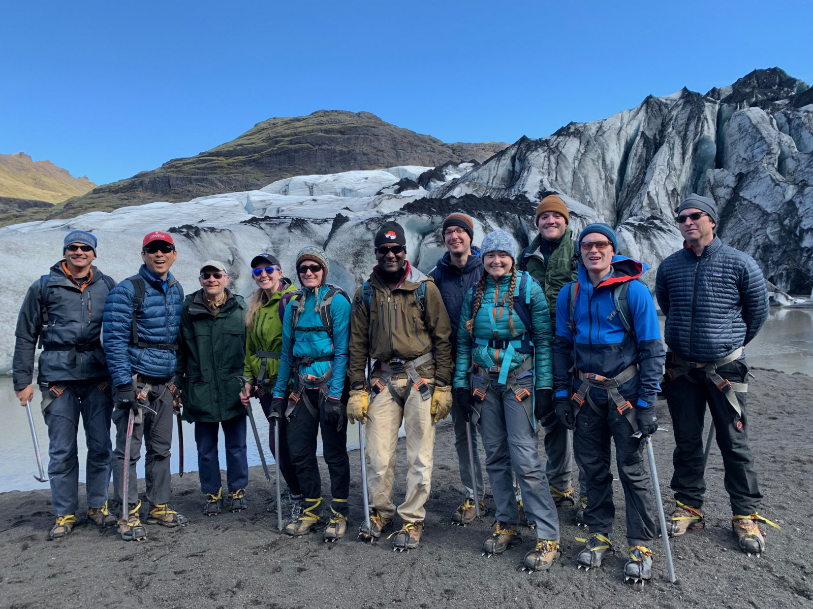 A group of climate researchers stand in front of a glacier in Iceland on a dark sandy patch of soil.
